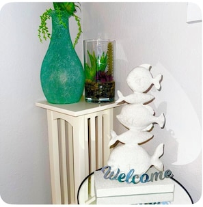 A teal vase with a plant, a glass container with rocks and plants, and a white fish-shaped decoration on a white console table.