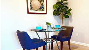 A dining area with a round glass table, two blue upholstered chairs, a potted plant, and a framed artwork on the wall.