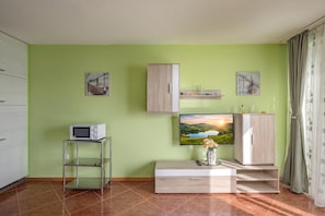 Una sala de estar con una televisión de pantalla plana montada en la pared, un mueble de madera, un pedestal de cristal para la televisión y un jarrón con flores sobre una mesa.