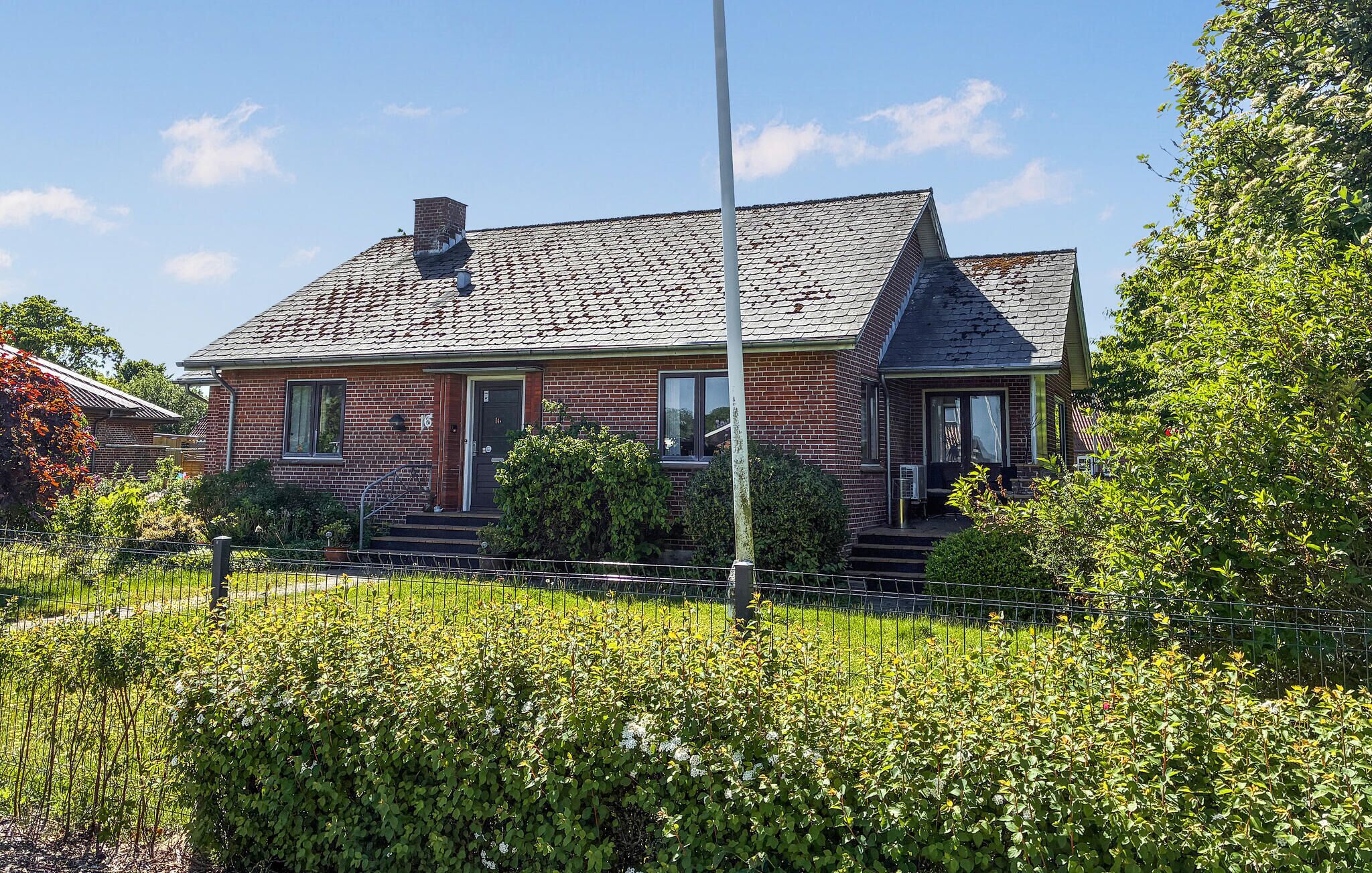 Une maison à deux étages, construite en briques, avec un toit à forte pente, un porche à l’avant et un jardin clôturé.