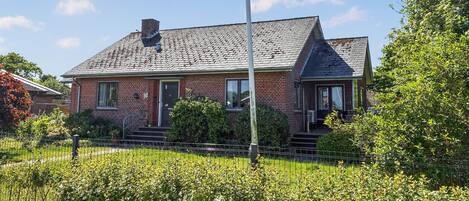 Une maison à deux étages, construite en briques, avec un toit à forte pente, un porche à l’avant et un jardin clôturé.