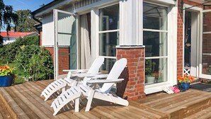 Una terraza de madera con tres sillas Adirondack blancas, un muro de ladrillo y ventanas amplias.