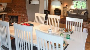 Un comedor con chimenea de ladrillo, sillas blancas, una mesa con comida y una sala de estar al fondo.