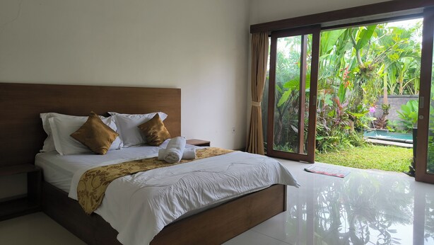 A modern bedroom with a large bed, wooden headboard, and a view of a lush garden through floor-to-ceiling windows. - Villa Patrisia Ubud (Abiansemal)