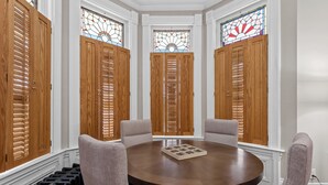 Una habitación con una mesa redonda, cuatro sillas y tres ventanas con vidrieras y contraventanas de madera.