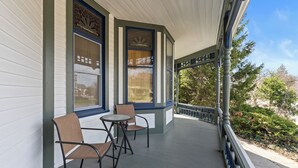 Un porche cubierto con dos sillas y una mesa, ventanas amplias con detalles decorativos, y vista a árboles y al cielo.