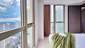 A hotel room with a large window, a bed with a green blanket, a door, and a view of the cityscape. - Aria By SanLiving - 2BR - Direct Mall (Jakarta)