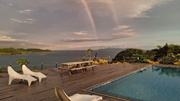 Ein Poolbereich mit Liegestühlen, einer Holzterrasse und Blick auf das Meer mit einem Regenbogen.