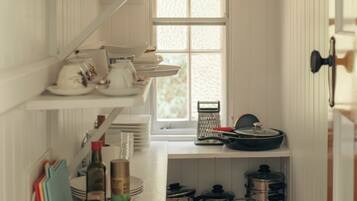 A narrow kitchen with white wooden walls, a window, and shelves holding kitchenware and utensils.