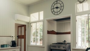 A kitchen with white wooden paneling, a stove, a clock, and a window with a view of greenery.