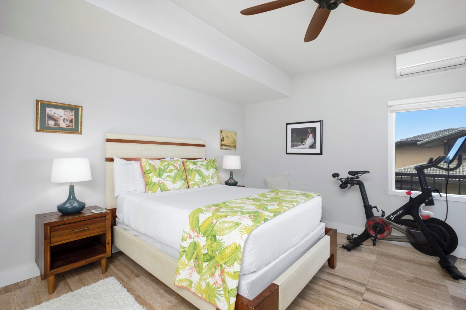 Une chambre à coucher avec un lit, deux tables de chevet, un ventilateur de plafond, un appareil de climatisation, un vélo et une fenêtre donnant sur un beau paysage.