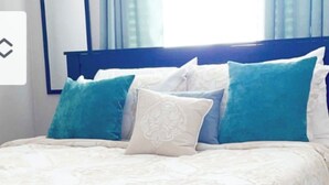 Una cama bien hecha con una colcha blanca y beige estampada, almohadas azules y blancas, y un cabecero de madera oscura.