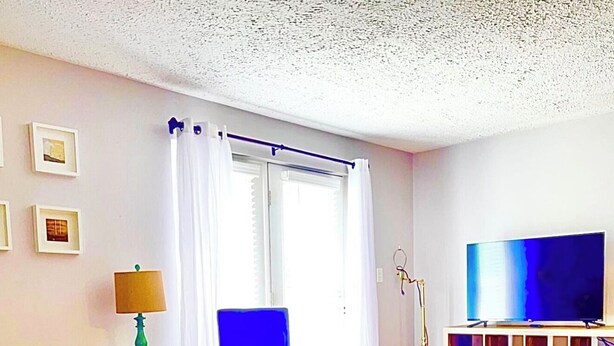 Un salón con una silla azul, un sofá amarillo, una mesa de centro redonda, un televisor sobre un mueble de madera y una lámpara de pie.