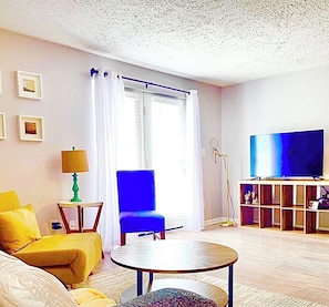 Un salon avec un fauteuil bleu, un canapé jaune, une table basse ronde, un téléviseur posé sur un meuble en bois et une lampe sur pied.