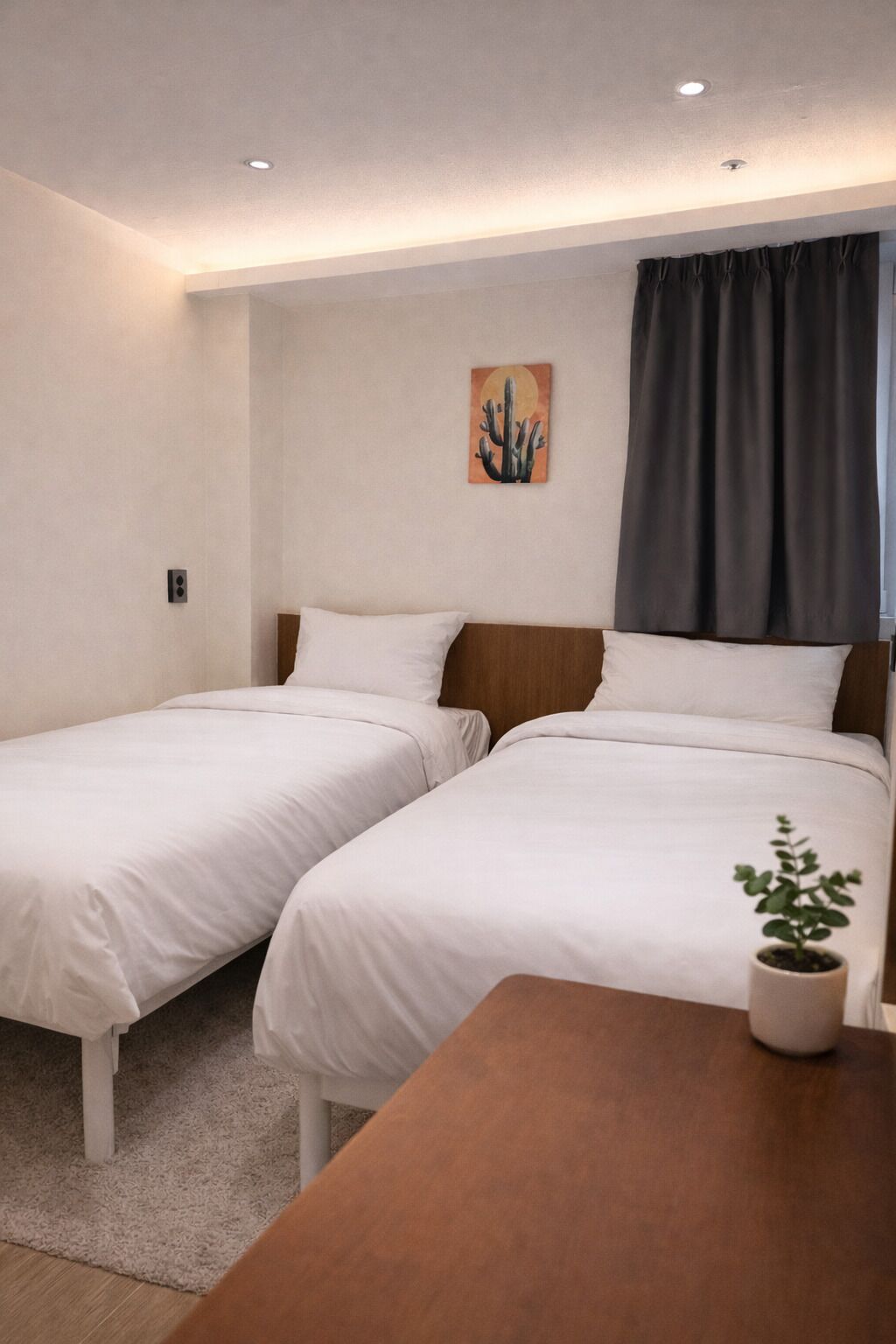 A hotel room with two single beds, a wooden headboard, a small table with a plant, and a painting of a cactus on the wall.
