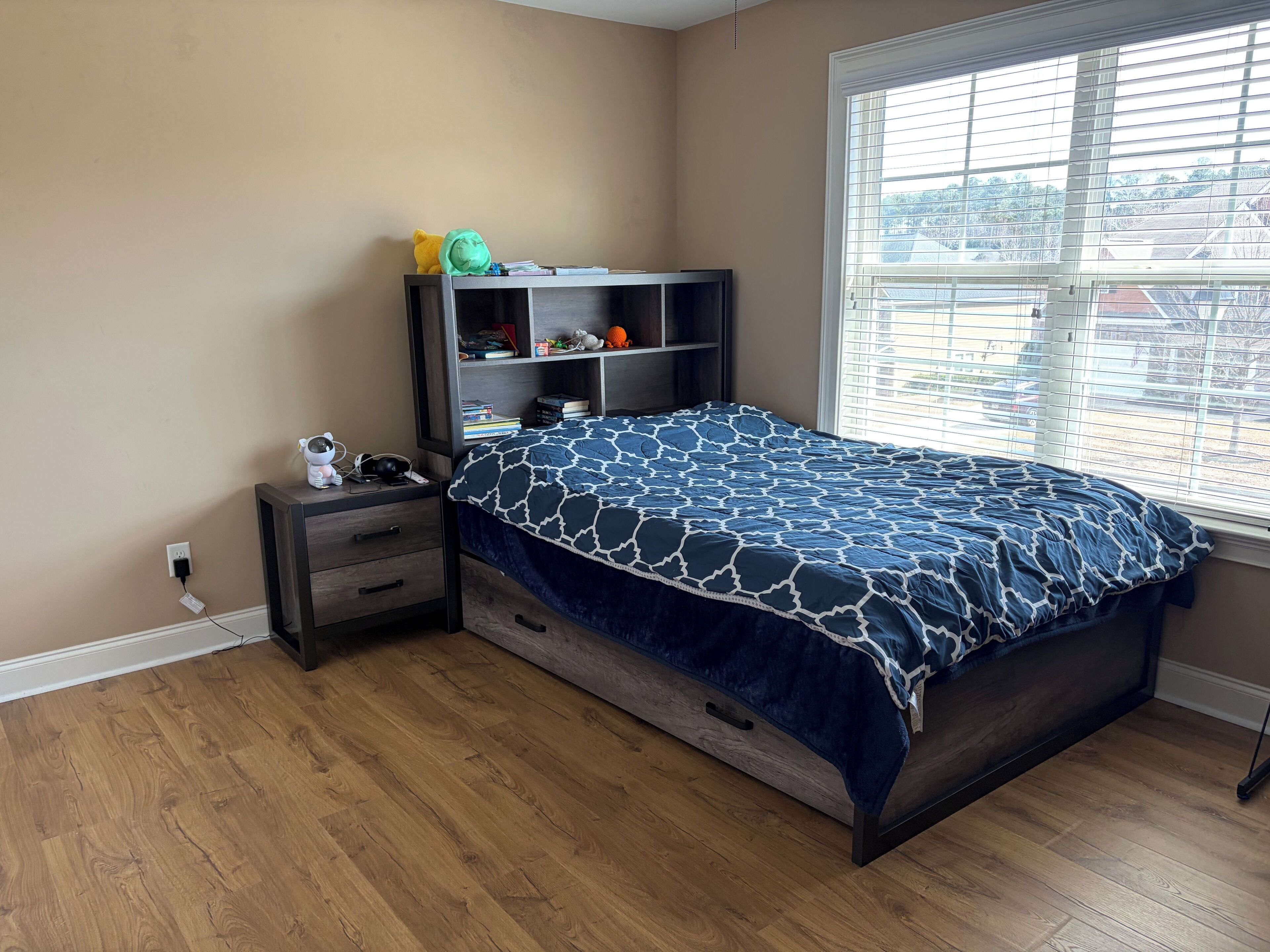 A bedroom with a bed, a nightstand, a bookshelf, and a window with blinds.