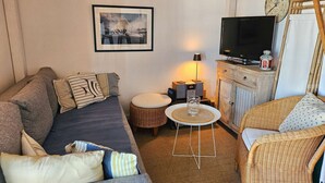 A cozy living room with a sofa, a wicker chair, a television, a clock, and a framed picture on the wall. - Beautiful 2-room apartment with Wi-Fi and mooring – View over the canals of Port Grimaud (PORT GRIMAUD)