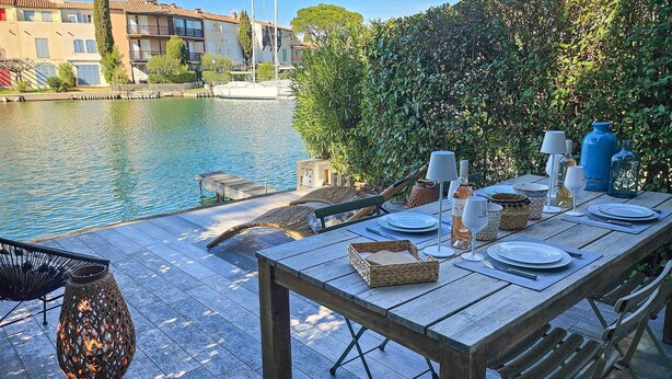 Un área de comedor al aire libre con una mesa preparada para dos, con vistas a un cuerpo de agua y una hilera de edificios.