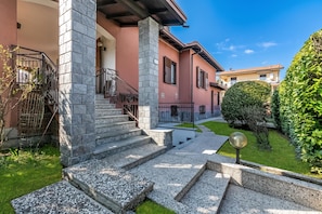 A residential building with a stone staircase leading to a porch, a well-maintained lawn, and a clear blue sky. - Villa Eva: Leggiuno 5-bed Villa for Families & Groups (Leggiuno)