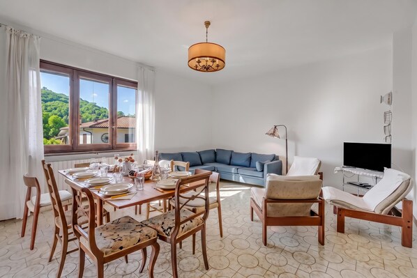 A well-lit living room with a dining area, a sofa, a TV, and a view of a green landscape through large windows. - Villa Eva: Leggiuno 5-bed Villa for Families & Groups (Leggiuno)