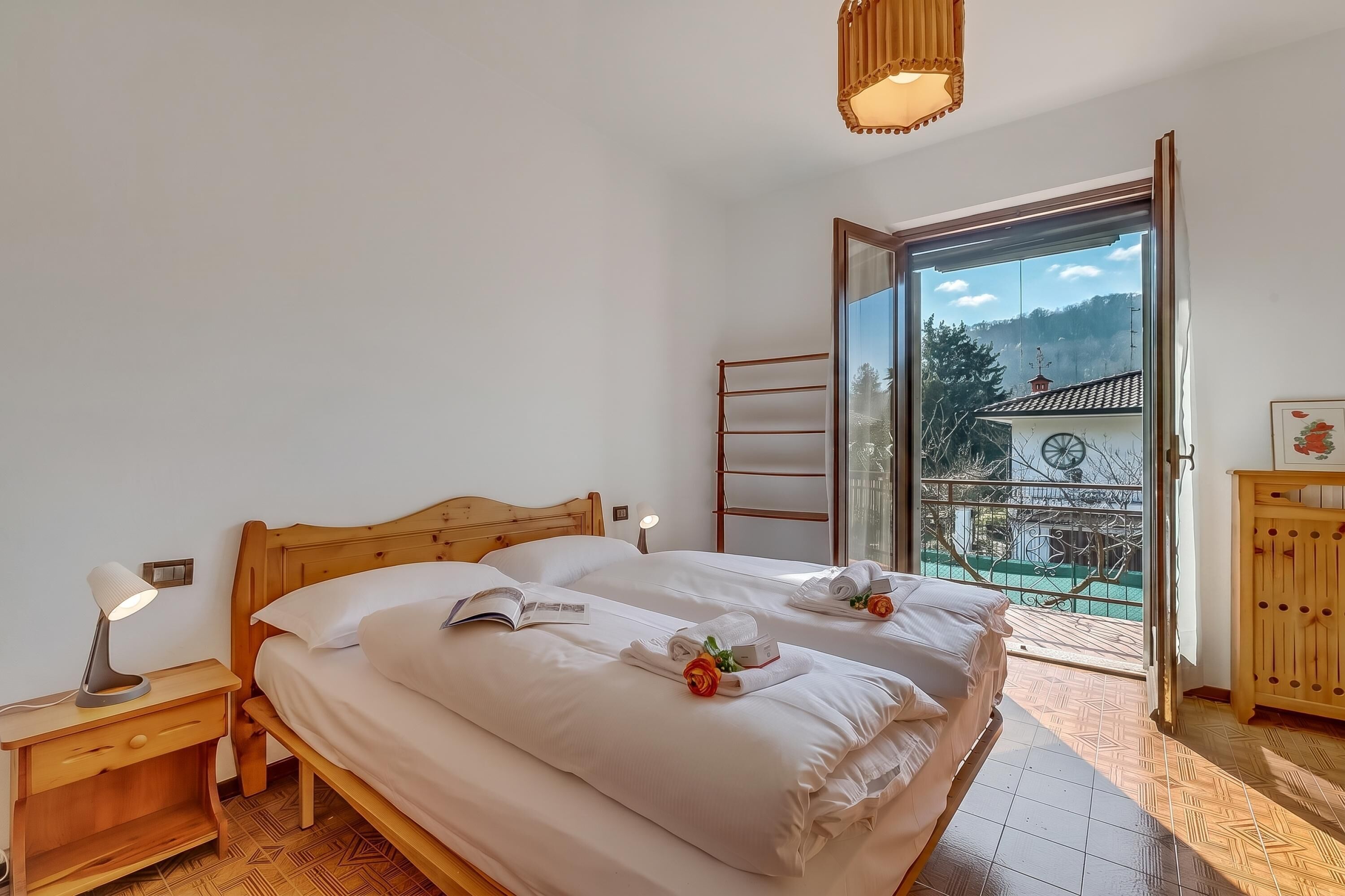 Una cama bien hecha con ropa blanca, un libro, una lámpara de noche, una mesita de madera, y una vista a un balcón con barandilla y una montaña al fondo.