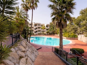 A swimming pool surrounded by palm trees and a stone wall, with a building in the background. - Apartment with pine forest views 800m beach (Salou)