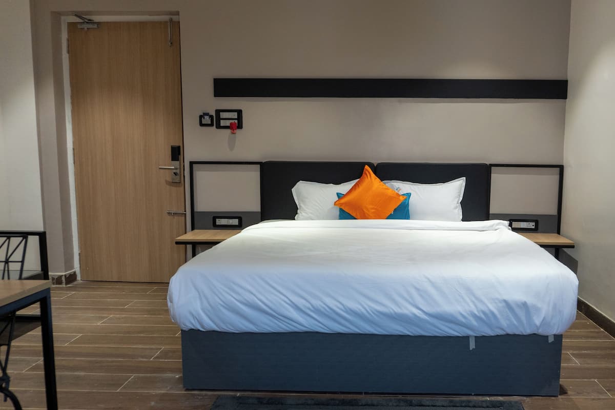 Una habitación de hotel moderna con una cama grande, una puerta de madera, una mesita pequeña y una mesita de noche con una lámpara.