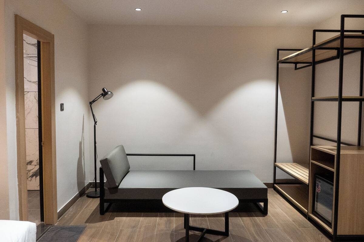 Una habitación pequeña con una cama, una mesita redonda, un sofá, una lámpara de pie y una estantería.