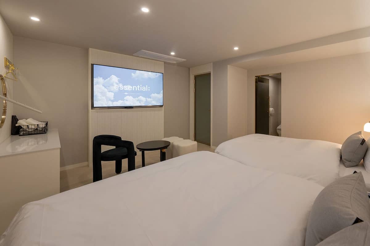 A hotel room with two beds, a TV screen displaying 'Essential,' a small table, a chair, and a bedside lamp.
