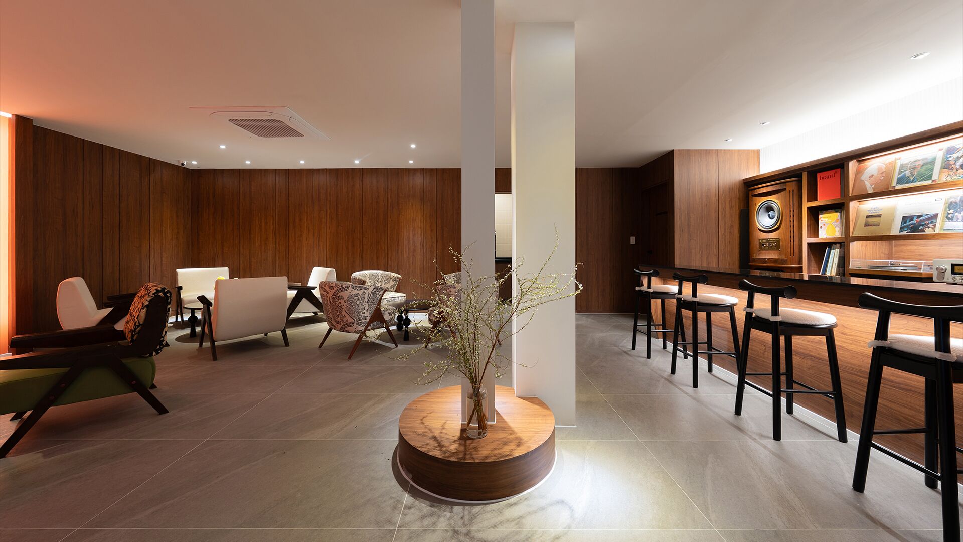 A hotel lobby with a reception desk, bar area, seating with chairs and sofas, a decorative vase with branches, and wooden paneling on the walls.