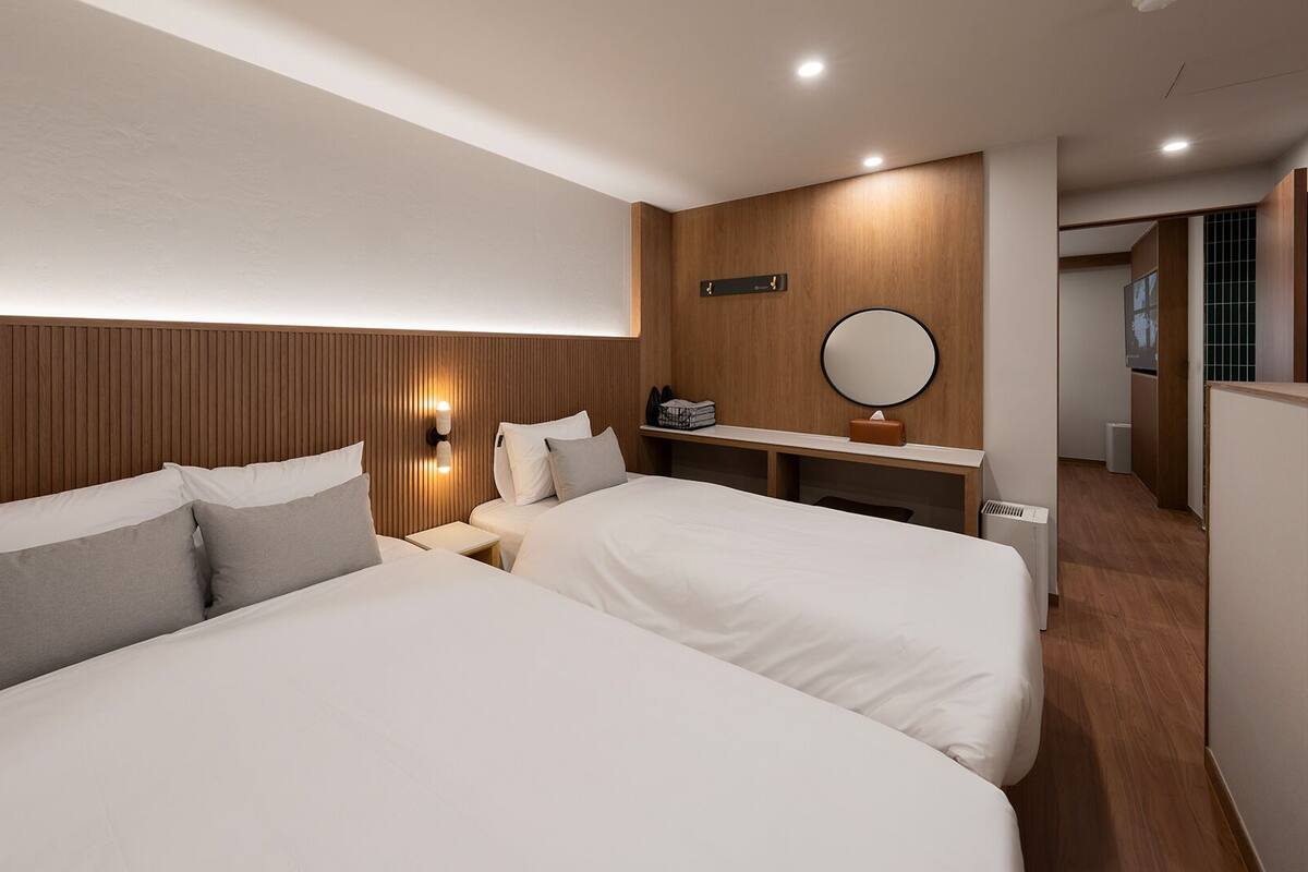 A hotel room with two beds, a wooden headboard, a bedside table with a lamp, a mirror, and a small shelf with a tissue box.