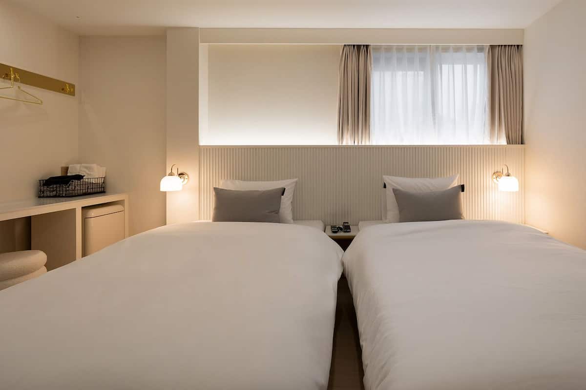 A hotel room with two beds, a bedside table, a desk, a chair, a hanging light fixture, and a window with curtains.