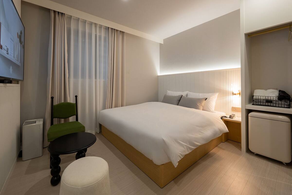 A hotel room with a bed, a chair, a small table, a TV, a desk with a lamp, and a mini-fridge.
