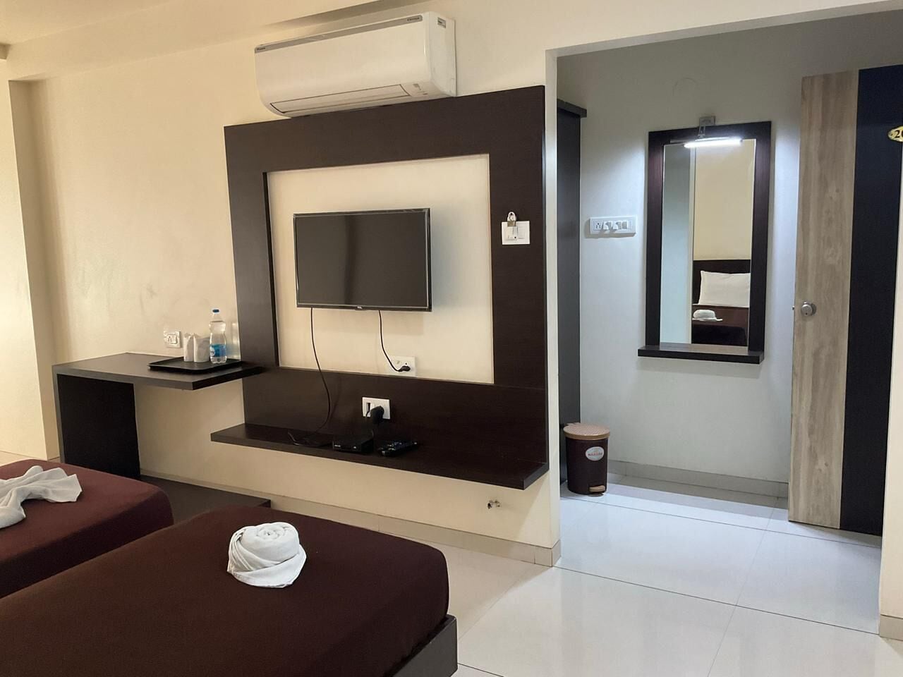 Une chambre d’hôtel avec un téléviseur à écran plat fixé à un mur foncé, une petite table avec une bouteille d’eau, une poubelle et deux lits avec des serviettes.