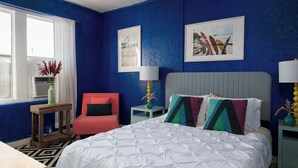 A bedroom with a large bed, a red chair, a window with white curtains, and two framed pictures on the wall.