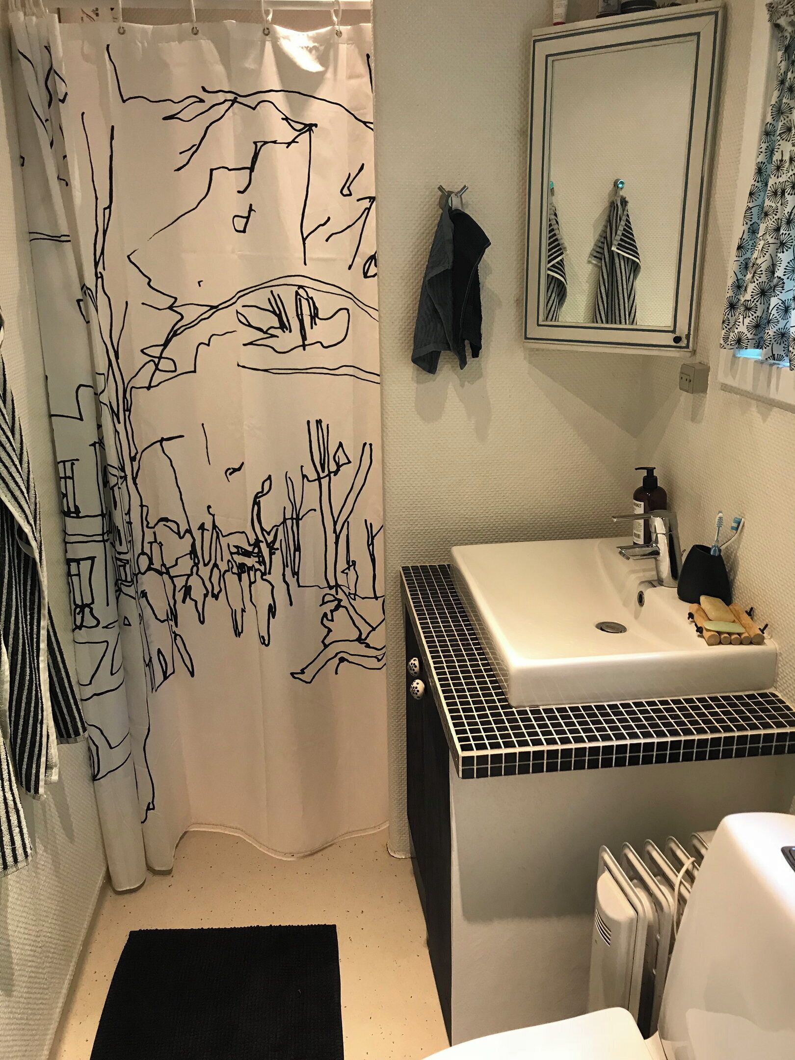 A bathroom with a black and white patterned shower curtain, a white sink with a black tiled base, a mirror above the sink, and a black bath mat on the floor.