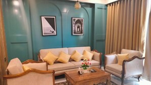 A living room with a teal wall, two framed pictures, a beige sofa with yellow cushions, a wooden coffee table, and a patterned rug. - HOTEL ABHIJIT (Uluberiya)