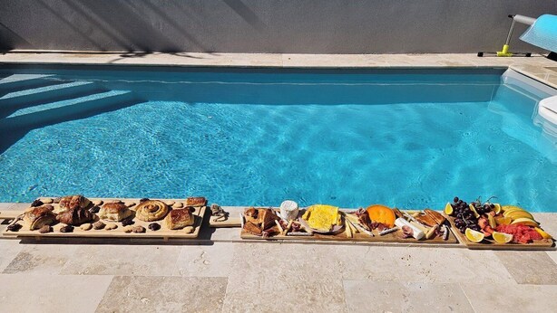 Area piscina con solarium piastrellato e un lungo tavolo di legno con un assortimento di cibo e bevande.