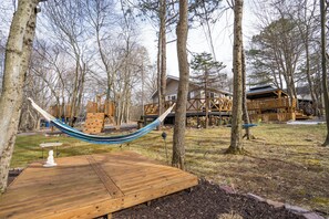 A wooden deck with a hammock, a play structure, and a house in a wooded area.