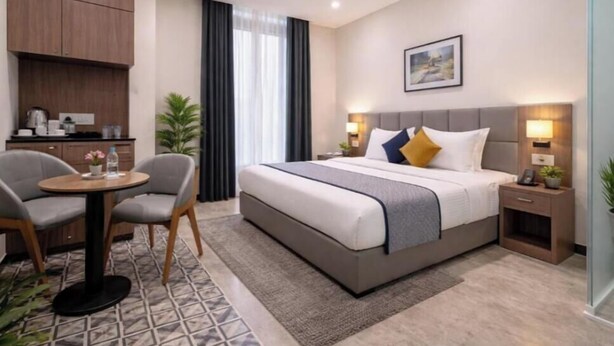 A hotel room with a bed, bedside tables, a desk with chairs, a small kitchen area, and a window with curtains. - Hotel Trawika (Ahmedabad)
