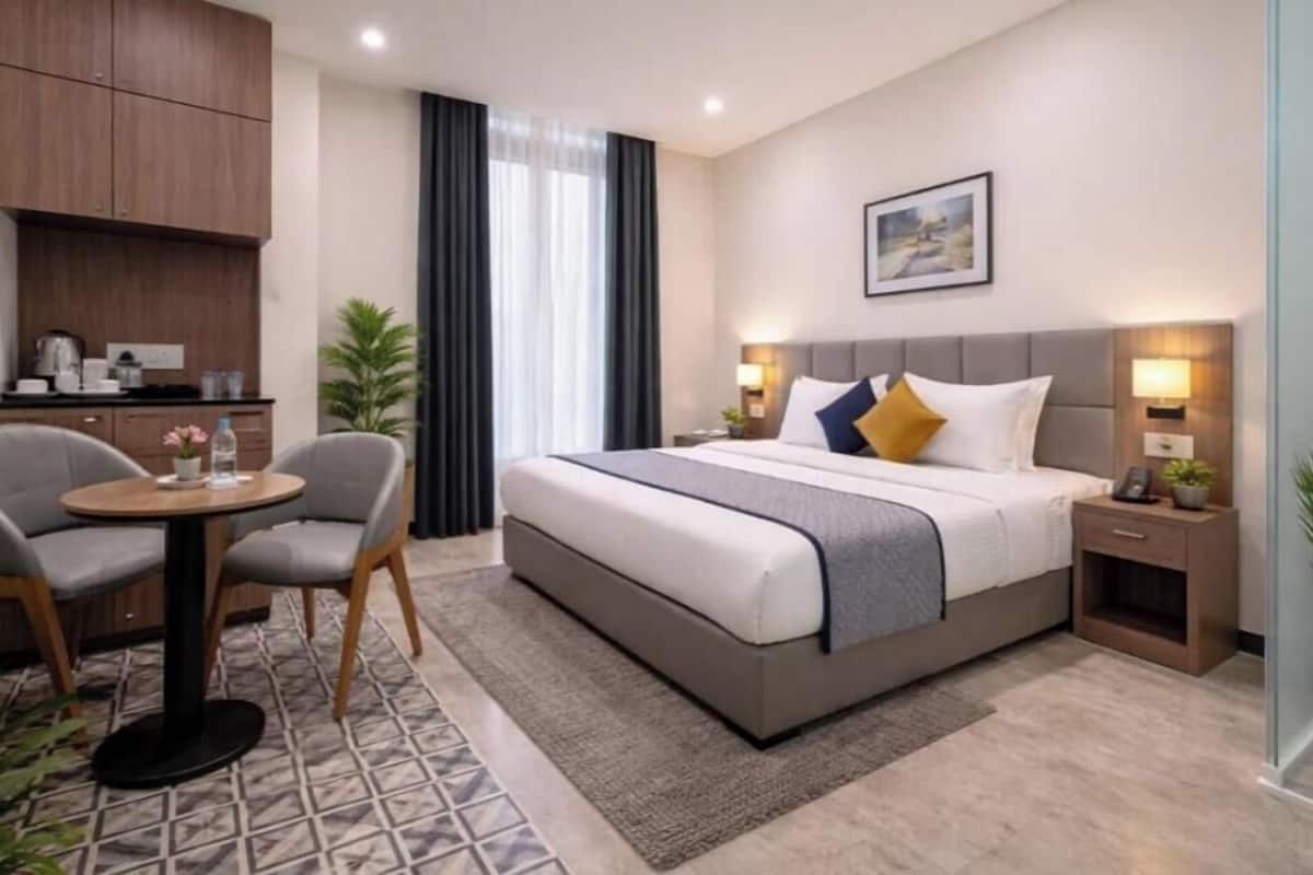 A hotel room with a bed, bedside tables, a desk with chairs, a small kitchen area, and a window with curtains.