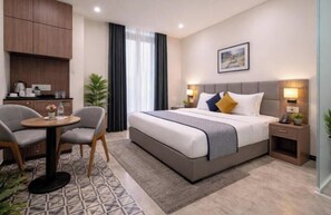 A hotel room with a bed, bedside tables, a desk with chairs, a small kitchen area, and a window with curtains. - Hotel Trawika (Ahmedabad)