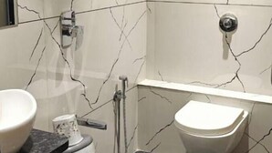 A bathroom with a white marble wall, a black floor, a white toilet, a white sink, and a shower head. - Hotel Trawika (Ahmedabad)