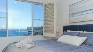A bedroom with a large window, a bed with blue bedding, a white throw blanket, and a painting of a beach scene.