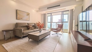 Una sala de estar moderna con un sofá, una mesa de centro y una silla. Hay un ventanal con cortinas, un cuadro en la pared y una pequeña mesa con un jarrón de flores.