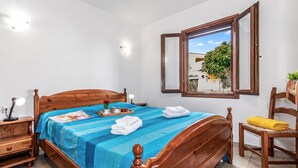 A bedroom with a wooden bed, a bedside table, a chair, a window with a view, and a wall-mounted light. - Le Case Della Cinta Hosue (San Teodoro)