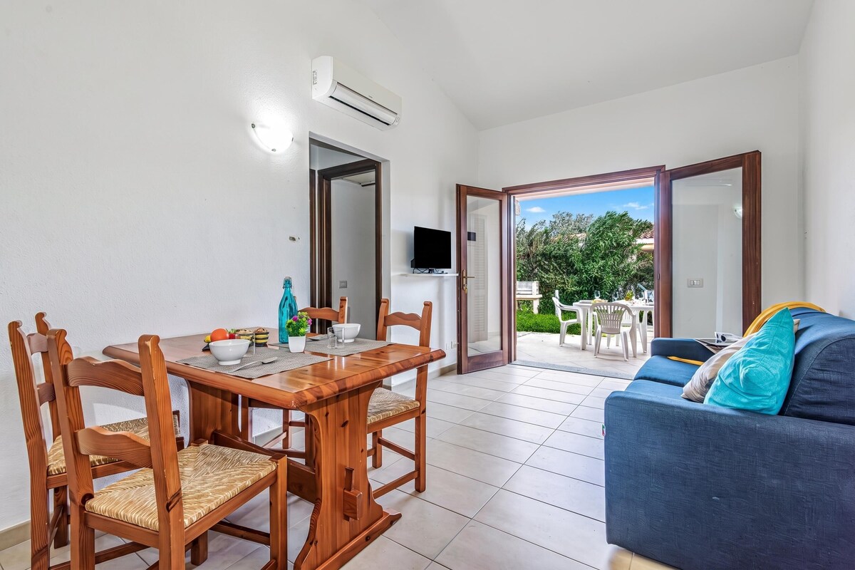 A dining area with a wooden table and chairs, a television, and an open door leading to an outdoor patio with a table and chairs.
