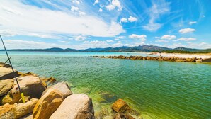 A rocky shoreline with clear turquoise water, a fishing rod, and distant mountains under a blue sky. - Le Case Della Cinta Hosue (San Teodoro)