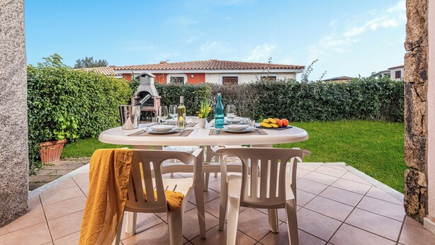A patio with a round table set for dining, surrounded by chairs, with a view of a house and a hedge in the background. - Le Case Della Cinta Hosue (San Teodoro)