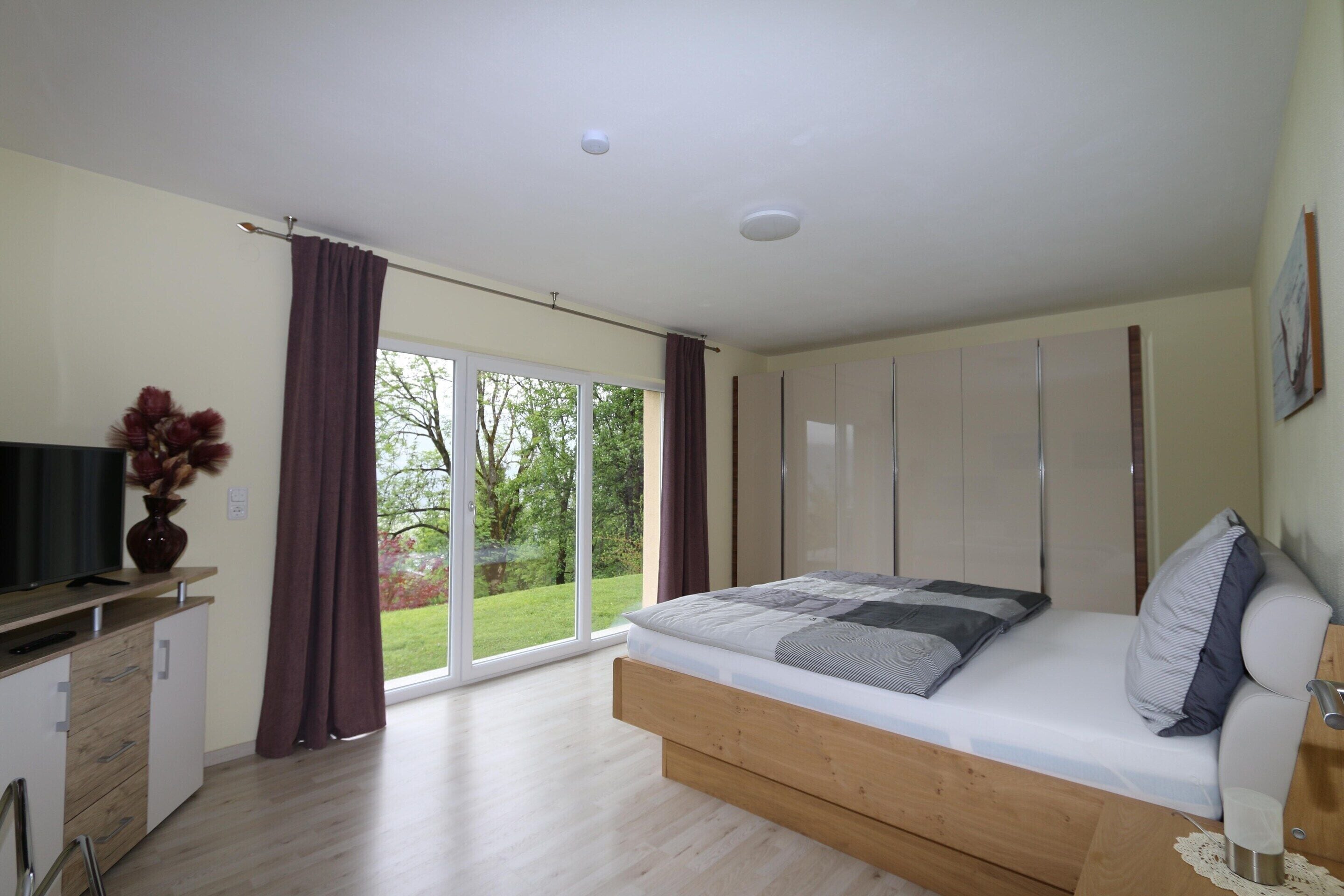 A bedroom with a bed, a desk, a TV, and a view of the outdoors.